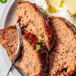 A fork taking a bite out of a slice of air fryer meatloaf on a plate with mashed potatoes and green beans.