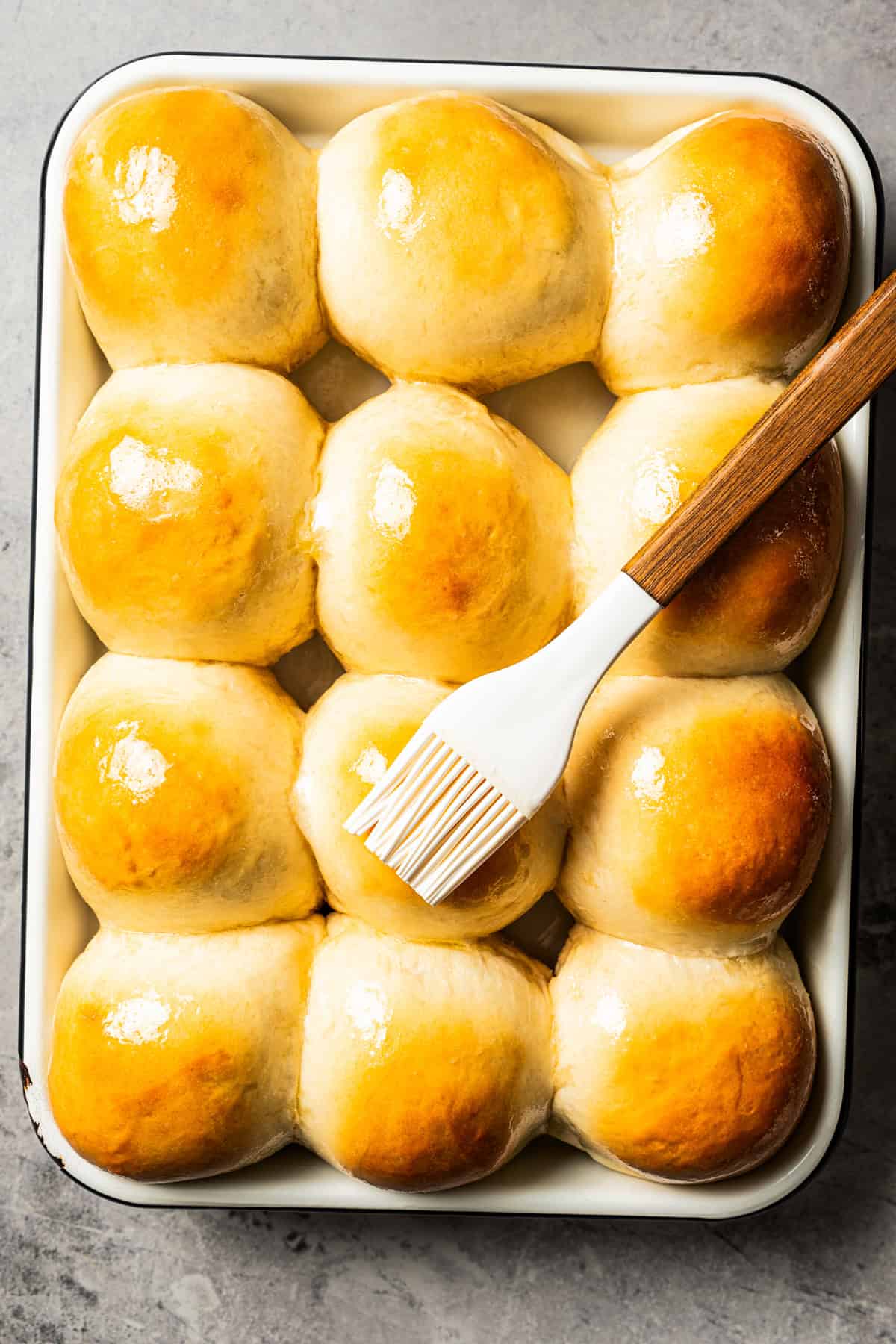 A basting brush is used to brush melted butter over freshly baked dinner rolls on a baking tray.