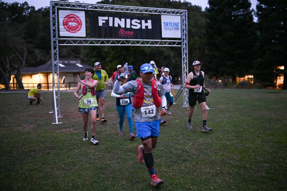 About ten trail runners starting a race on a lawn, just in front of a temporary metal arch with logo text for Scena Performance & Skyline 50k, and a large FINISH white text on black background banner across the top, park buildings in the background, and trees behind them.