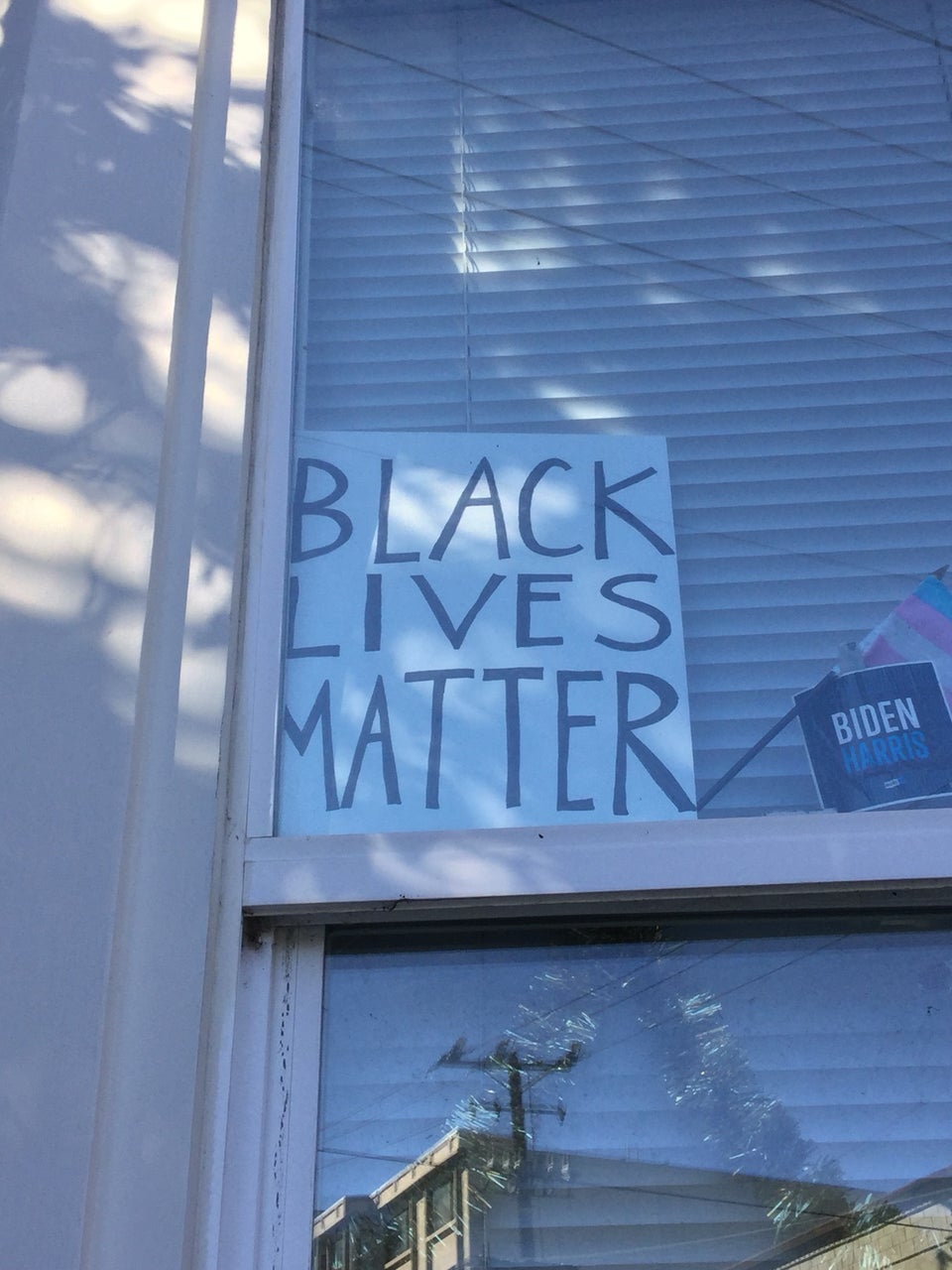 Hand drawn BLACK LIVES MATTER sign, black marker on white cardboard, inside the window of a building, mini-blinds closed behind it holding it up, a small BIDEN HARRIS sign next to it.