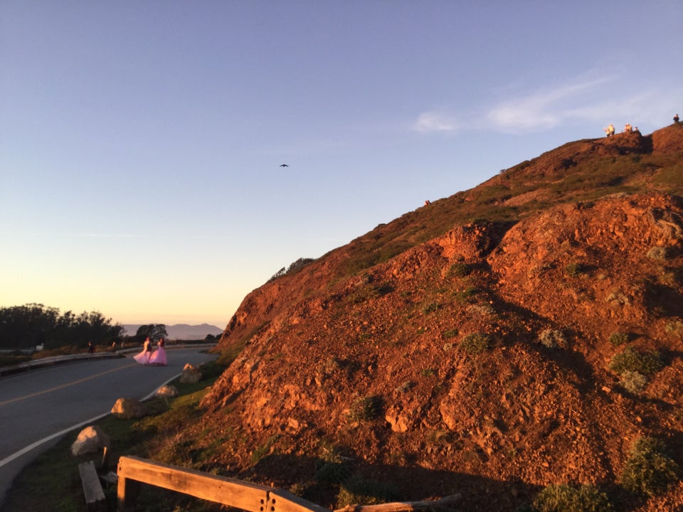 Large mostly dirt and rocks hill in front lit up with orange light from the sunset, a road off to the left, with two women in pink dresses in the distance walking down the middle of the road.