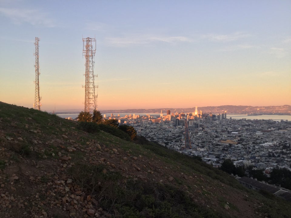 Twin Peaks towers, downtown San Francisco in the distance, looking down Market Street from above, its streetlamps lit, the sun glinting off the taller downtown skyscrapers.