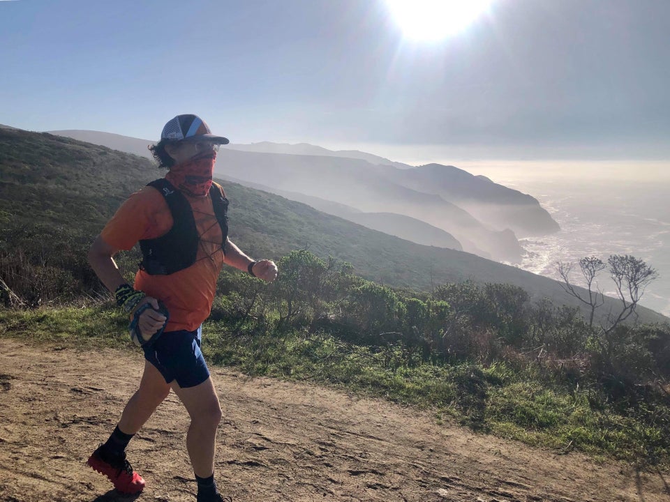 Tantek running downhill on a trail with mountains, hills, and an ocean shore in the distant background, under a sunny blue sky.