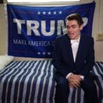 Nick Fuentes sitting on a bed with a Trump flag behind him answering questions during an interview.