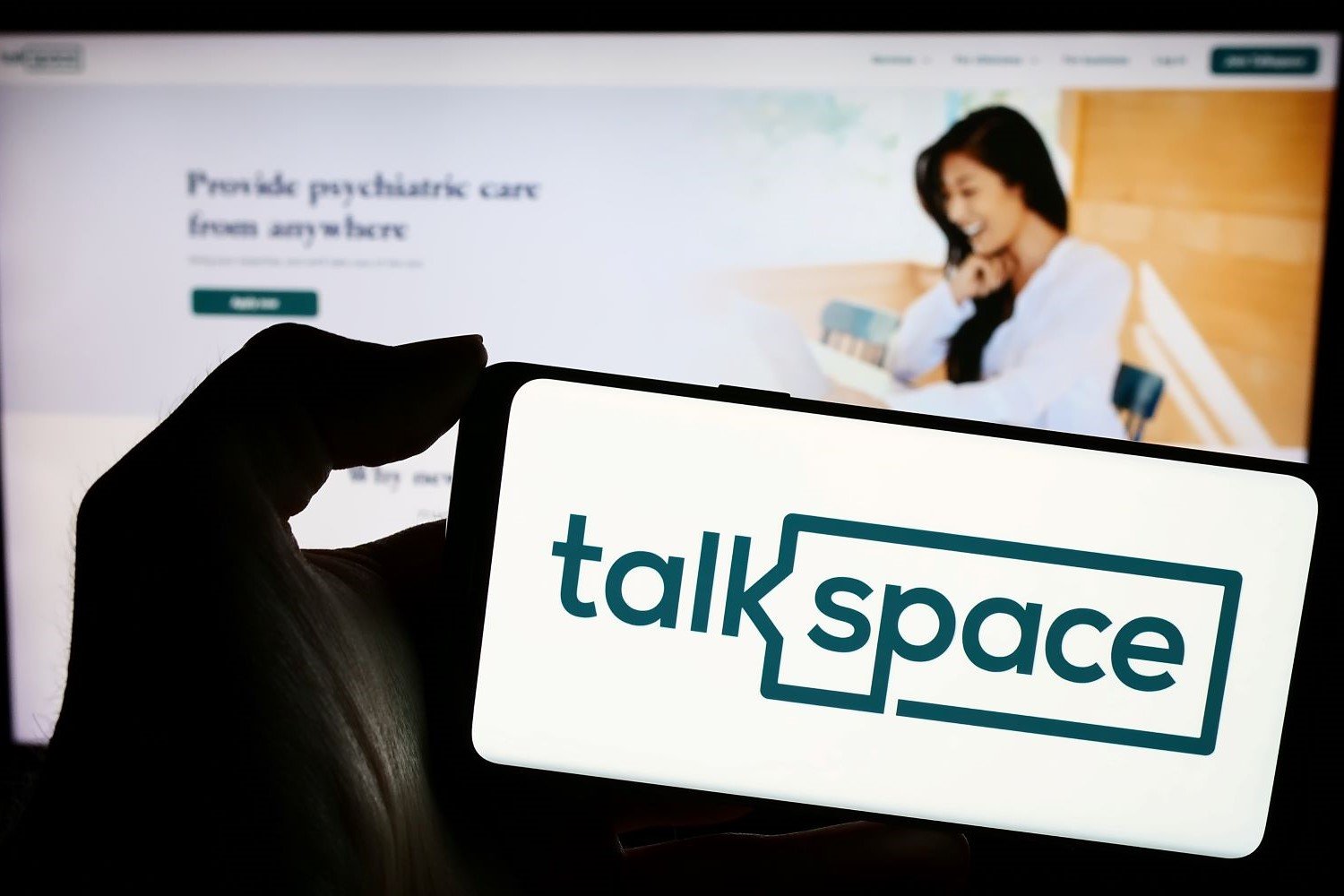 A phone displaying the Talkspace logo in front of a computer screen showing the company's website.