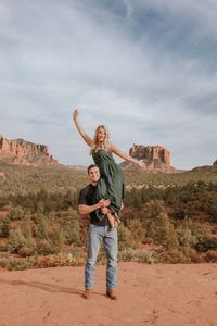 The couple is wearing dark and earthy tone colors for their outfits in the summer at Arizona. She is wearing a dress and wedges. He is wearing a black shirt, jeans and brown shoes.