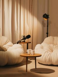 Comfy, cozy podcast studio generated with Midjourney AI has Call Her Daddy hosted by Alex Cooper podcast set vibes. Inviting podcast living room set up, podcast microphones, comfy cozy beige aesthetic chairs, modern minimalist interior design style