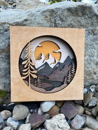 "Introducing our Rustic Wood Layered Mountain Scene, a stunning laser cut artwork that brings nature's beauty to your home. Each piece is carefully stained, resulting in a unique creation every time. Bring the mountains home. Order your Rustic Wood Layered Mountain Scene now and embrace nature's wonder. 9\" square. 6 layers of 1/4' Maple wood."