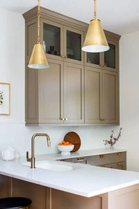 a refined taupe kitchen with shaker cabinets mostly lower and a couple of upper ones, white stone countertops and a backsplash, gold pendant lamps