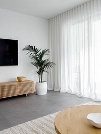 Dark and dull grey living area transformed into a warm and inviting space. The white linen sher curtains, oak side table, seagrass pouf, soft ivory rug, oak entertainment unit and indoor palm tree balance the cold of the grey floor tiles. Click now to see more photos of this beautiful living room