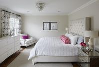 Beautiful bedroom features walls painted tan, Benjamin Moore Olympic Mountains, lined with a cream linen tufted wingback bed dressed in white bedding and red print bolster pillow flanked by chevron mirrored nightstands, Regina Andrew Furniture Chevron Antique Mirror Small Chests, and tan ceramic lamps illuminated by a Robert Abbey Bling Flush Mount facing a white curved 9 drawer dresser.