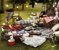 A twilight picnic setting with textiles, lights, baskets, food and drinks in a park surrounded by grass and trees.