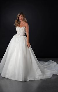 Elevating timeless bridal elegance, this designer ballgown wedding dress marries classic charm with contemporary flair. Featuring a scooped strapless neckline to introduce a corset bodice with a unique embroidered floral lace and exposed boning. The back showcases a classic tulle tie-up detail, while a detachable oversized bow accents the waistline, leaving its long lacy streamers to dramatically cascade down the full length of the wedding gown. From the timeless Basque waist, a gathered ballgown skirt billows in luxe organza, offering a clean and modern contrast to the romantic embellished bodice. This strapless ballgown wedding dress surprises once more with the addition of discreet hand pockets, perfecting the balance of sophistication and convenience.