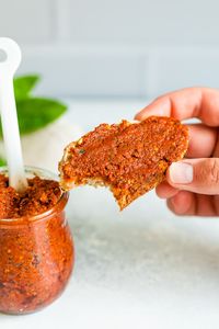 Hand holding a slice of bread spread with sun-dried tomato pesto. The bread has a bite taken out of it. There is a jar and spoon full of sun-dried tomato pesto.