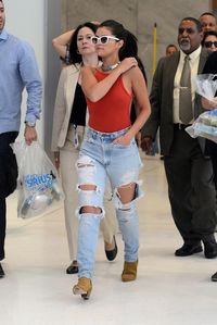 Selena Gomez wears white sunglasses, a red bodysuit, distressed denim, and tan suede boots