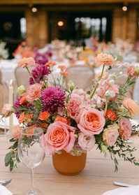 Lush arrangement features soft pink roses, magenta dahlias, and coral carnations, complemented with delicate greenery in a terracotta pot. Rustic elegance for a charming outdoor table setting.