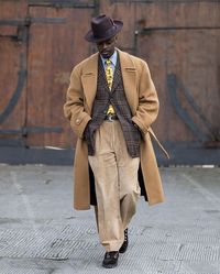 Kevis Manzi in a stetson hat, a big camel coat, a blazer, a shirt and tie, wide corduroys, and hiking boots.