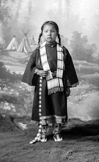 Full length studio portrait of Dakota girl, wearing long necklace and belt, standing in front of painted backdrop. Date: 188? Creator: D. F. Barry. David F. Barry Collection. Denver Public Library Special Collections, B - 42.