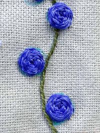 A close-up photo of blue flowers made with a woven wheel stitch using 6 strands of embroidery floss. The green stem is stitched with perle cotton using a stem stitch. The beige Charles Craft Monaco Needlework Fabric is in the background, and you can see some of the blue marks drawn with the UNIQUE Sewing 2-in-1 Dual-Tip Wash-out/Air Erasable Fine Tip Marking Pen around the edge of the blue flowers; DMC Embroidery Floss, DMC Cotton Perle Thread, DMC Satin Floss 8 Pack - Radiant Treasures, DMC Lig