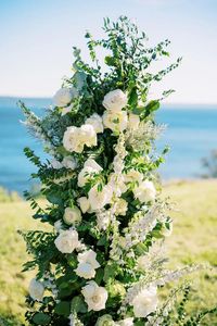 Upright column ceremony arch designed with greenery, white roses, and delphinium. A perfect outdoor summer wedding ceremony with a stunning view of the water.