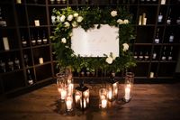 A wedding welcome sign surrounded with greenery and white roses makes a beautiful frame with candles and lanterns at the base.