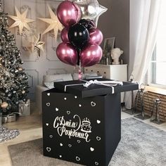 a black present box with pink and silver balloons in front of a decorated christmas tree