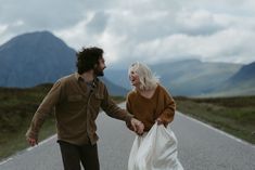 a man and woman holding hands while walking down the middle of an empty road with mountains in the background