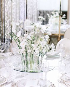 white flowers are in a clear glass vase on a dining room table set with silverware