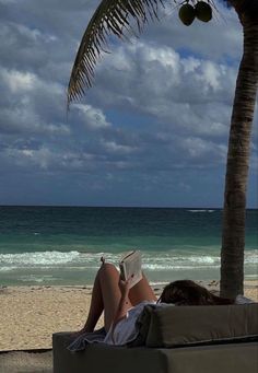 a woman laying on top of a couch under a palm tree next to the ocean