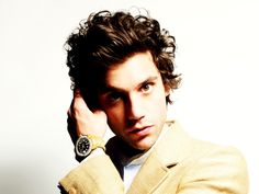 a man with curly hair wearing a tan suit and watch on his left hand, posing for the camera