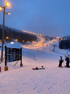 people on skis and snowboards at the bottom of a snowy slope with lights in the background