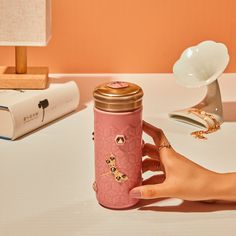 a hand holding a pink coffee mug on top of a white table next to a lamp