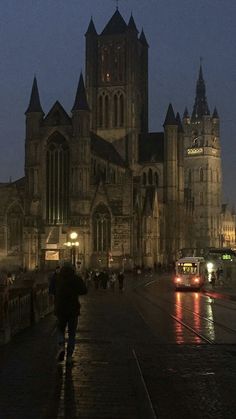 people are walking on the sidewalk in front of an old church at night with traffic passing by