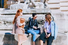 three women are sitting on a stone wall talking to each other and one is holding a book