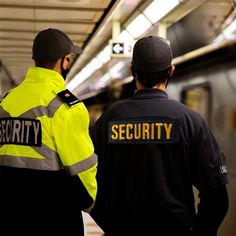 two security officers standing next to each other in a subway station, looking at the train