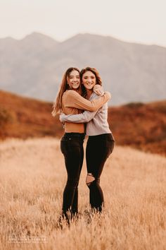 two young women hugging each other in a field with mountains in the backgroud