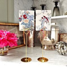 two gold candlesticks with birds on them sitting on a marble counter top next to pink flowers