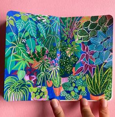 a hand holding up an open notebook with plants on the cover and pink wall behind it