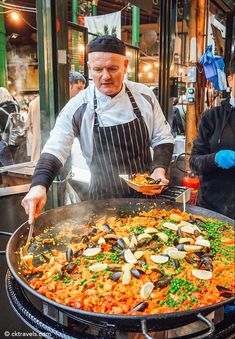 a man in an apron is cooking food on a large skillet while another man looks on