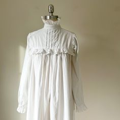 This beautiful long white nightgown evokes the Victorian era with its feminine and delicate details. Made from soft, breathable 100% cotton, it features long sleeves with ruffled cuffs and a high collar trimmed in eyelet lace. It gathers at the yoke which is also trimmed in white eyelet lace, creating a timeless vintage silhouette. The back fastens with three white buttons for an old-world charm.  Expertly sewn, Victorian nightgown. Treat yourself to a historic experience or delight someone who Cottagecore White Nightgown For Home, White Cottagecore Nightgown For Home, White Cottagecore Nightgown, Feminine Long Sleeve Cotton Nightgown, Cottagecore Ruffled Nightgown For Daywear, White Cotton Long Sleeve Nightgown, Feminine Long Sleeve Ruffled Sleepwear, Long Sleeve Cotton Nightgown With Lace Trim, Cotton Long Sleeve Nightgown With Lace Trim