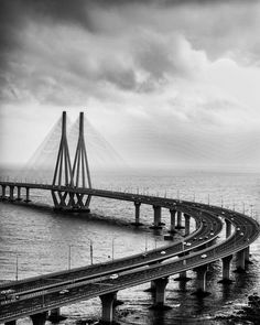 a black and white photo of a bridge over the ocean with cars driving on it