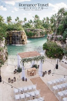 an outdoor ceremony setup with white chairs and greenery on the ground, in front of a waterfall