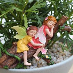 two little fairy figurines are sitting on a branch in a potted plant