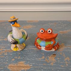 two ceramic figurines are sitting on a wooden table, one has a crab and the other is a frog