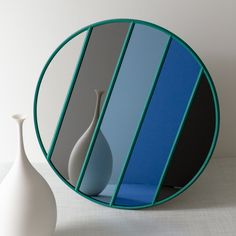 a white vase sitting next to a blue and black striped mirror on a table top