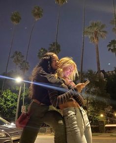 two women hugging each other in front of palm trees at night with their cell phones