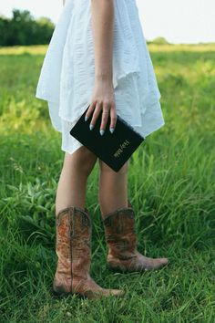 a woman standing in the grass with her boots on, holding a bible and wearing cowboy boots