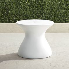 a white vase sitting on top of a cement floor next to a green hedge covered wall