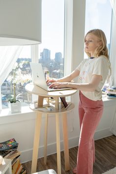 a woman standing in front of a laptop computer on a wooden table next to a window