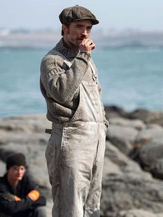 a man in overalls standing on rocks next to the ocean with another man sitting down